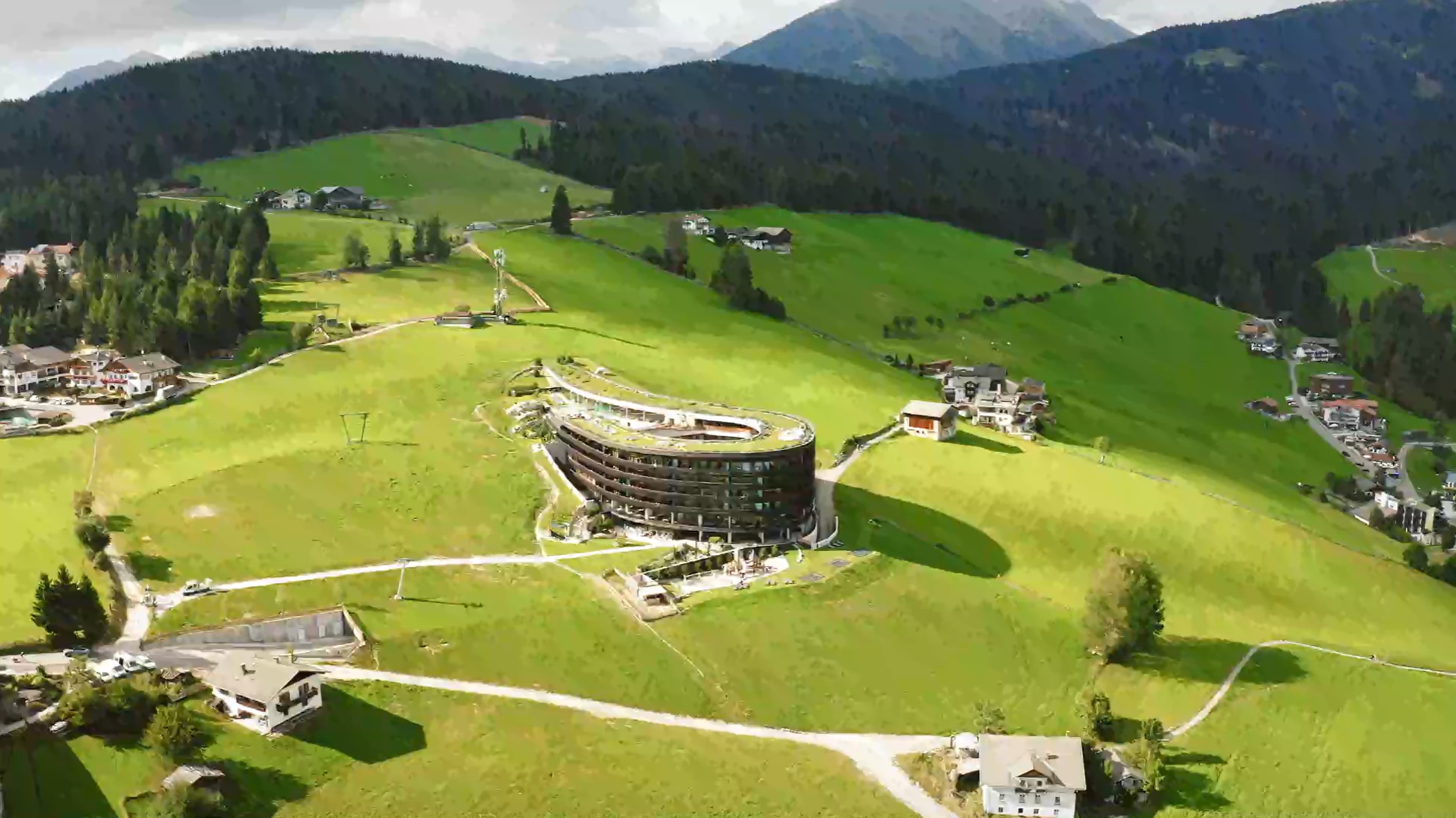 Luftaufnahme eines modernen, ovalen Gebäudes in grüner, hügeliger Landschaft mit verstreuten Häusern und Bergen im Hintergrund. - Plugintests